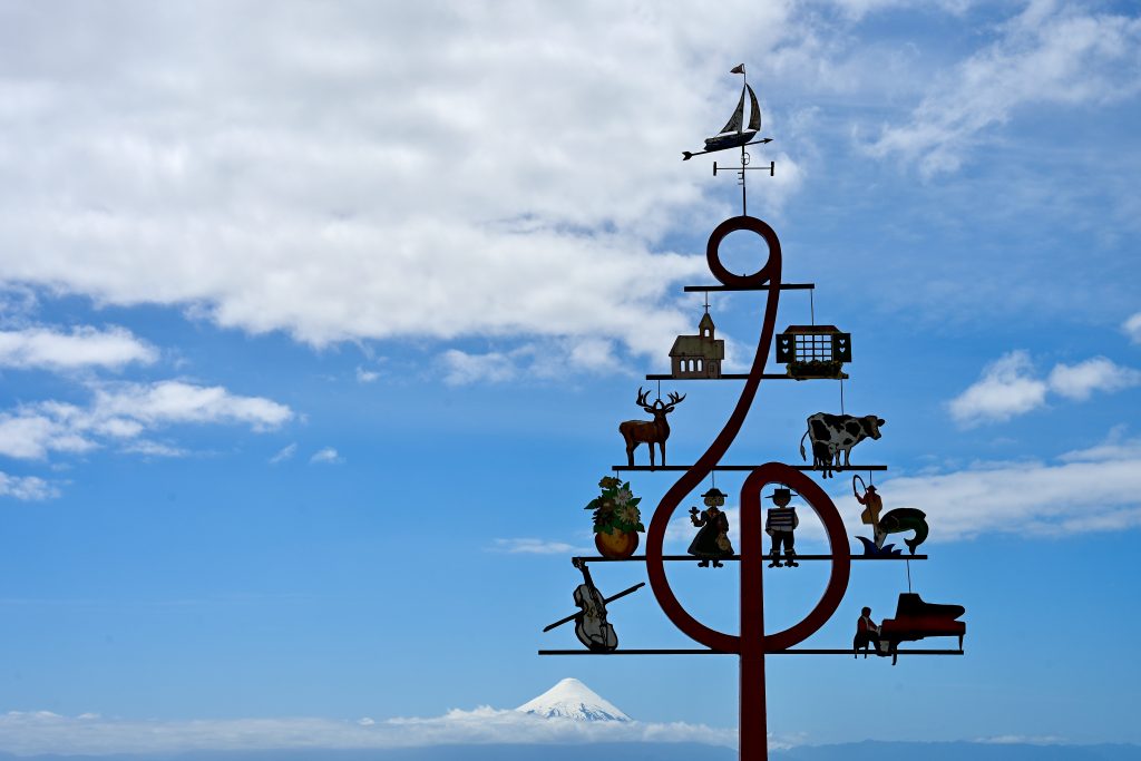 A blue sky with white clouds in the background. To the right hand side is a silhouette of a sculpture in the same of a fir tree with symbols relating to the town. Just noticeable in the centre bottom is a snowcapped volcano