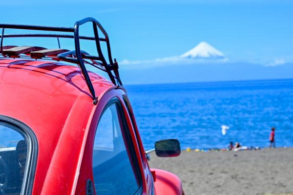 Half of a red Beetle car to the left hand side which is parked overlooking a beach and lake with a snow-capped volcano on the horizon. Pale blue sky above