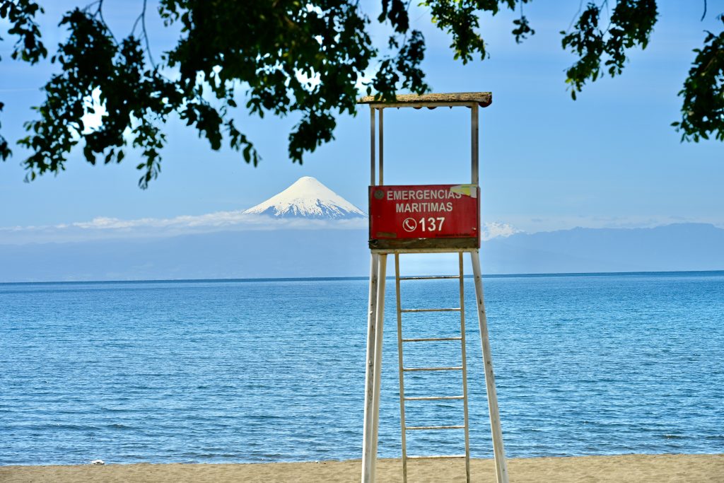 Row of sand at the bottom of the frame, with lake above. On the horizon the snowcapped cone of a volcano against a pale blue sky. In the foreground a red lifeguard seat on tall white ladder and legs. Tree branches above