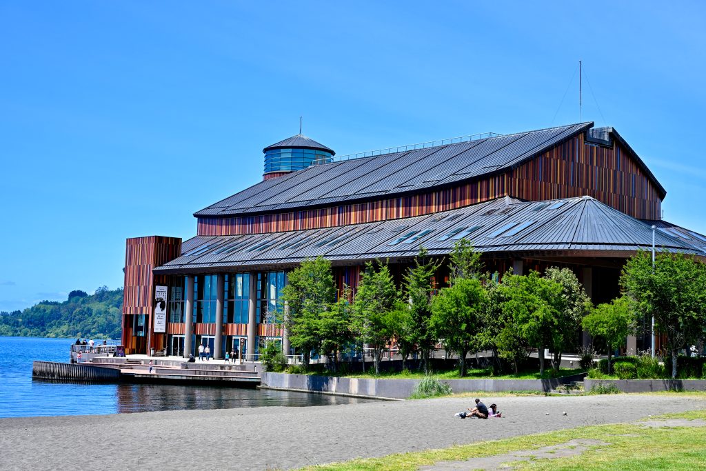 A wooden building, shaped a little like a boat, Sts on the edge of a lake. It appears to have striped wood underneath its roof. Blue sky above