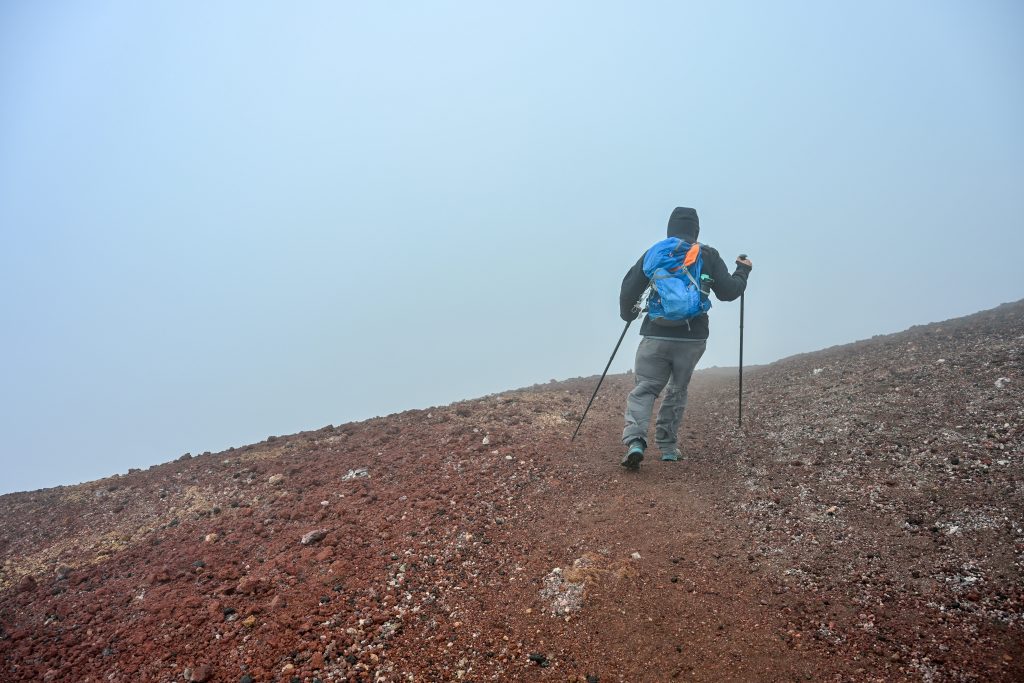 A man walks with poles away from the camera up a hill of dark red earth towards a wall of cloud. He is wearing a cagoule with hood up, a blue backpack and grey trousers