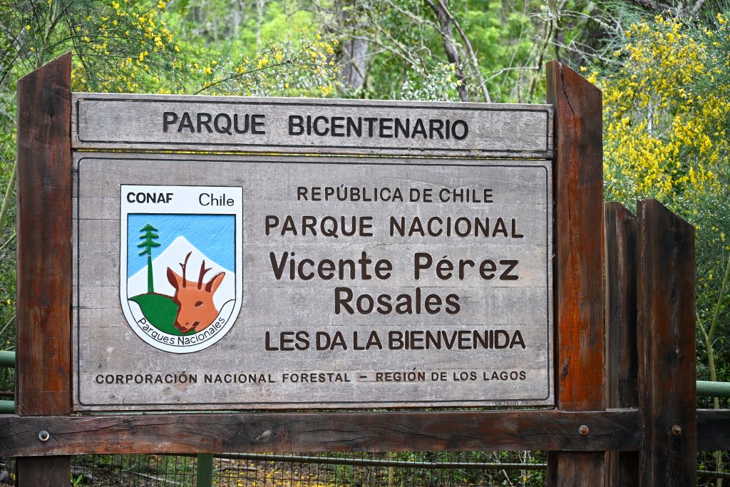 A large wooden sign fills the frame. It advertises Parque Nacional Vincente Perez Rosales with a shield to the left of the sigh featuring a volcano, monkey pule tree and deer. Trees behind.