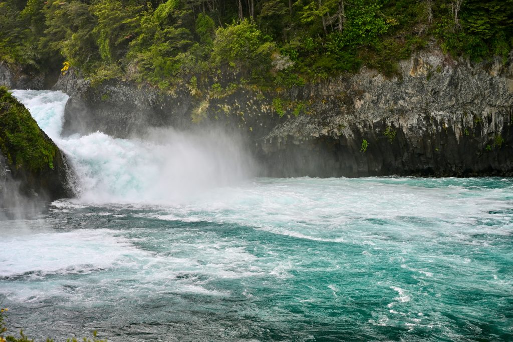 Icy blue water across the bottom of the frame. Dark rocks on the far side with trees on top. Water comes down a water fall to the left into the river. It is frothy white.