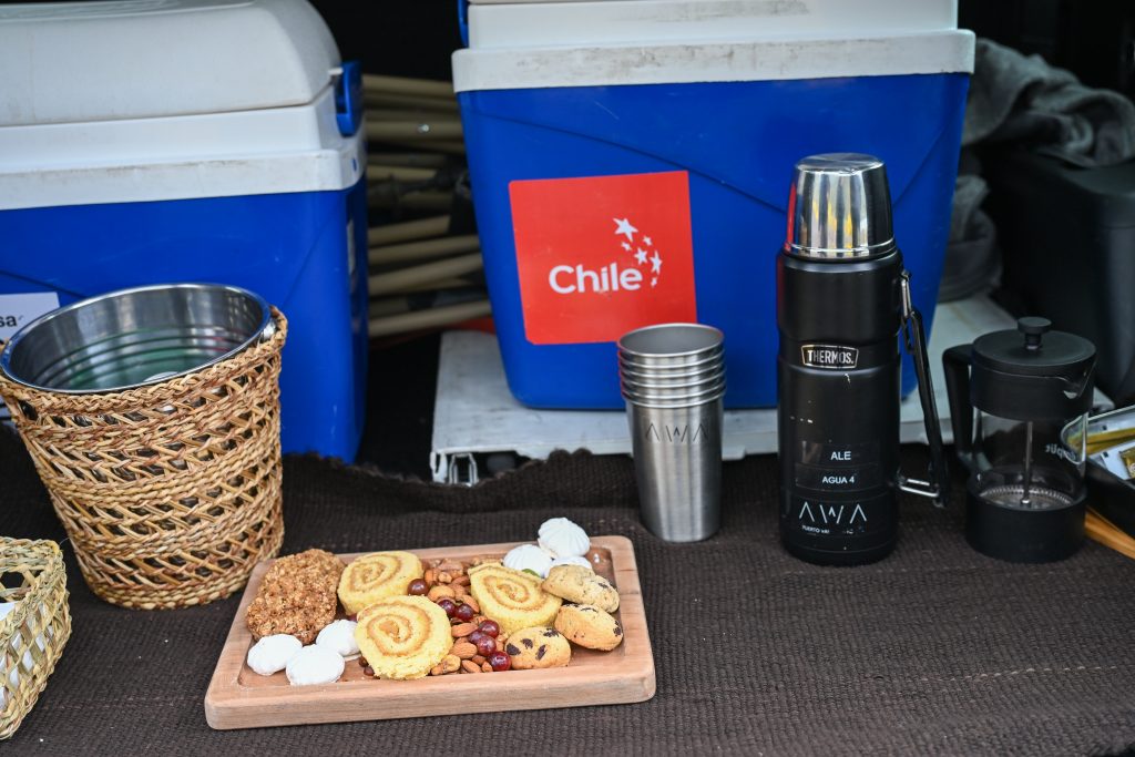 View into a car boot with two blue and white freezer boxes at the back. One has a red Chile sticker on it. In front is a black flask, silver cups, wicker bin and a wooden plate filed with biscuits, cakes and nuts