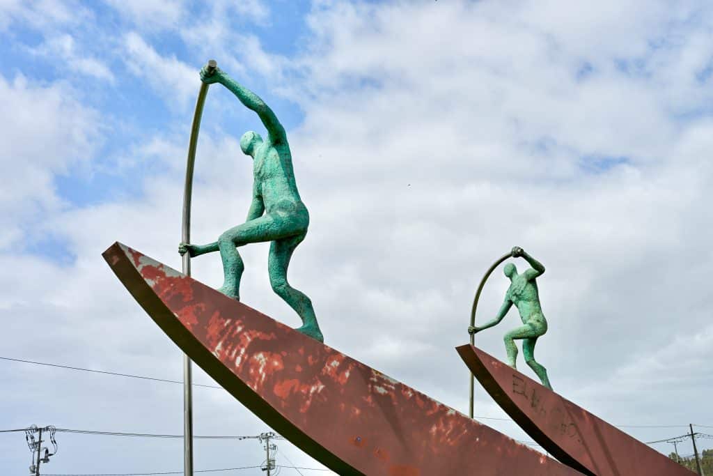 A metal sculpture enters the frame from bottom right. It forms two brass boat shapes, each with a turquoise male figure standing on the boat with an oar as if paddling