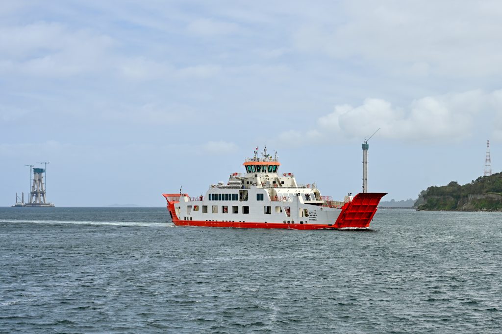 A white car ferry with red ramp at front and back, held in upwards position, makes its way across a grey body of water. Green cliff to the right. Grey sky above