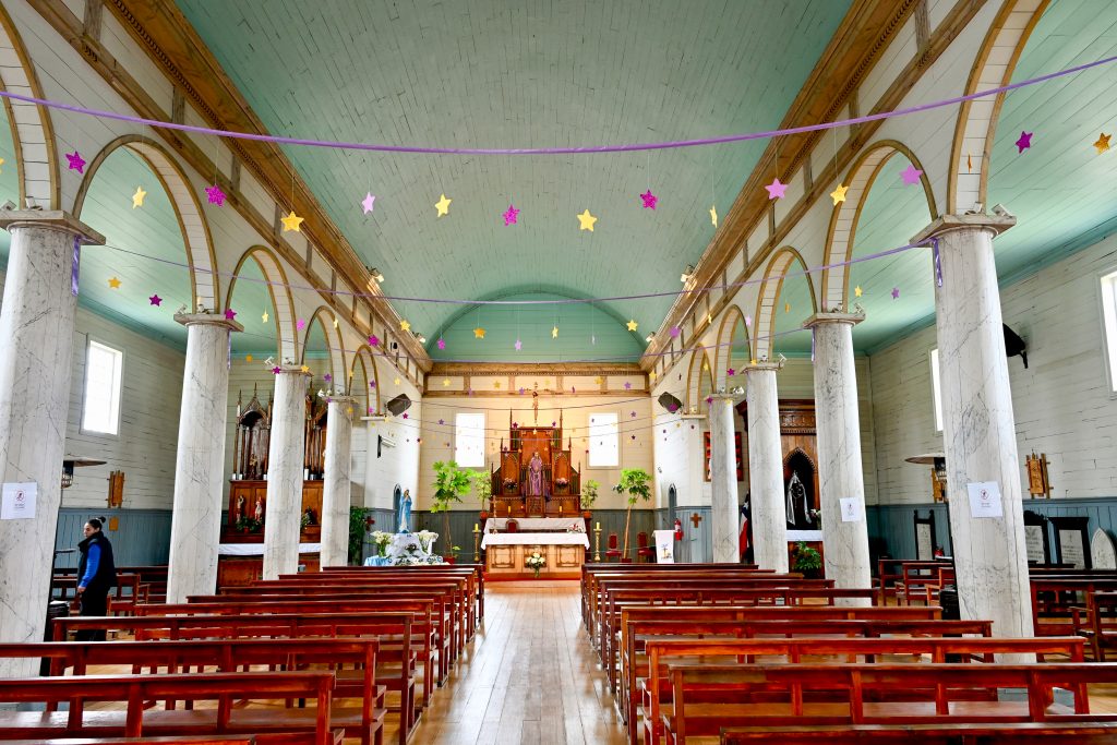 Looking down the aisle of a beautiful old church, simply decorated with white arched pillars and a. turquoise ceiling. Rows of wooden pews. A wooden alter carving at the far end. Strings of yellow and purple starts across the ceiling