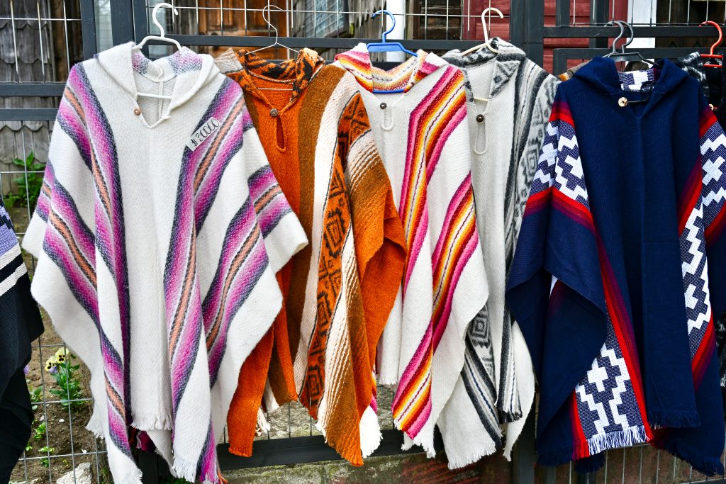 A line of 5 woven ponchos hang on coat hangers from a wire fence. The ponchos are different colours - white and purple, orange and white, white and pink, white and grey, and navy blue stripes.