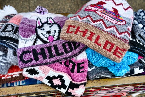 A pile of knitted hats sits on a table. They feature the word Chiloe and have pictures of huskies and boats on them in pinks, purples beige and grey