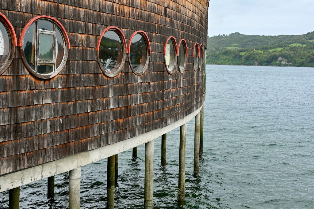 A circular wooden building on stilts stands over the water with trees on the horizon. The building has a row of circular port hole style windows