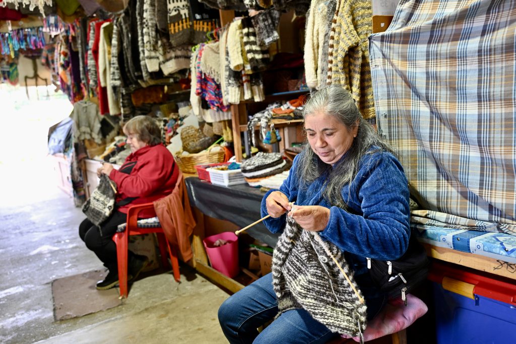 Two ladies sit in front of market stalls of textiles knitting jumpers. The lady on the left wears and red coat, and the lady to the right wears blue coat