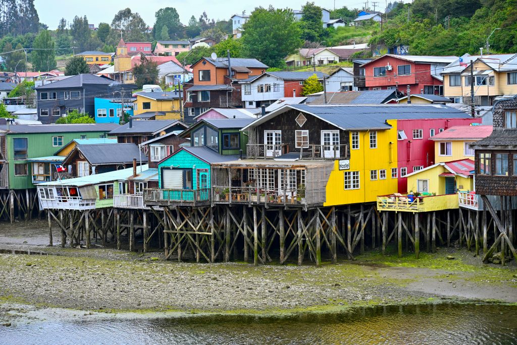 A row of multicoloured wooden huts on stilts stand on a muddy sandbank with water in front. The stilt houses cover the hill behind.