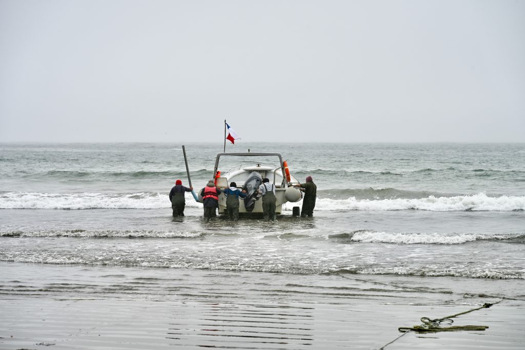 5 men push a white boat out into the waves of a very green sea. Grey sky above