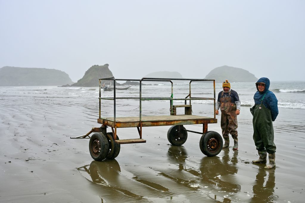 A metal trolley on wheels stands on a wet sandy beach in the mist and rain. Lumpy hills in the background. Two men in waterproof trousers and cagoules stand to the right of the trailer