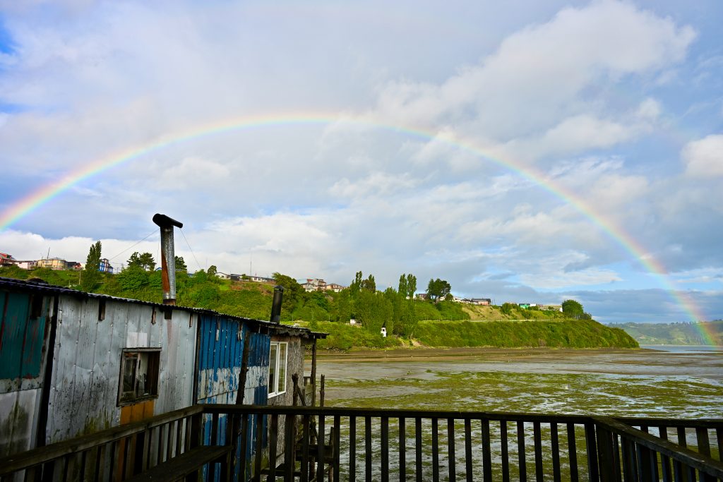 A beautiful half rainbow stretches across the width of the frame. In the foreground are the railings of house with a wooden shack to the left. In the horizon are green trees and houses on a hillside. muddy river bed in-between