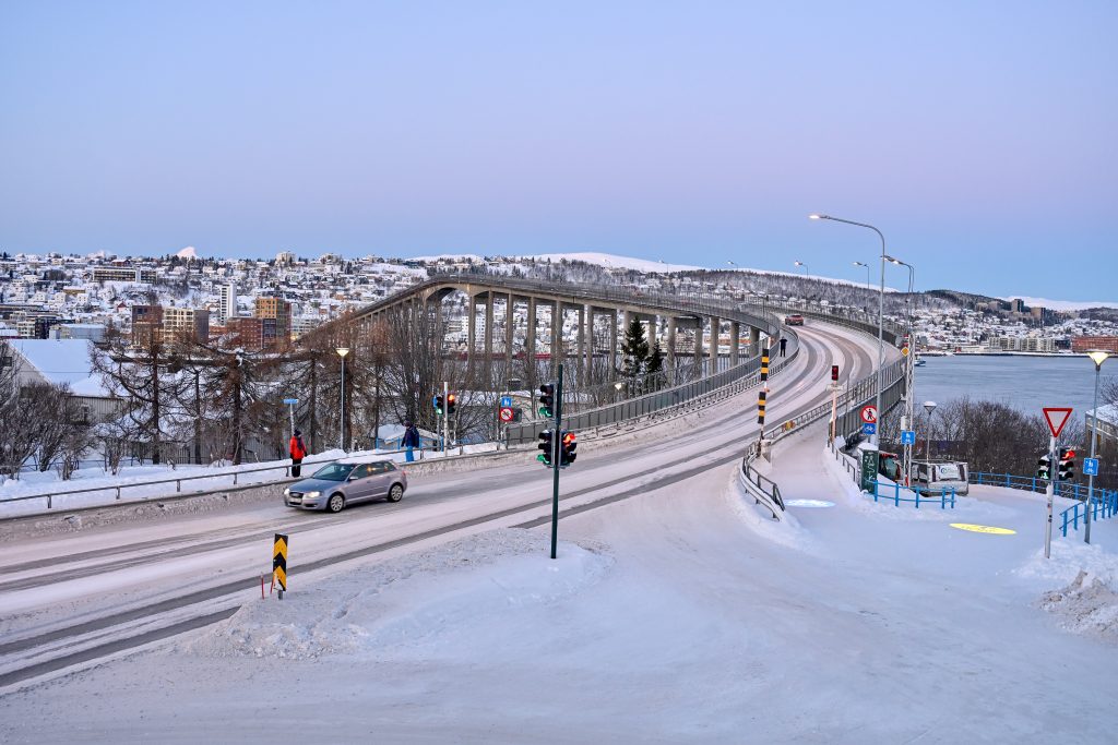 View across a snowy road bridge to snow covered land and buildings behind. Pale grey sky above. Car driving off the bridge to the bottom left