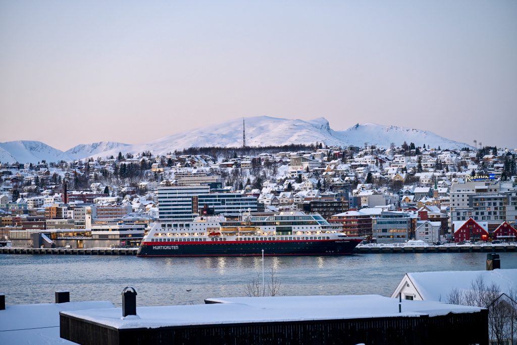 A large ferry ship sits on water in front of a town with snowy mountain behind