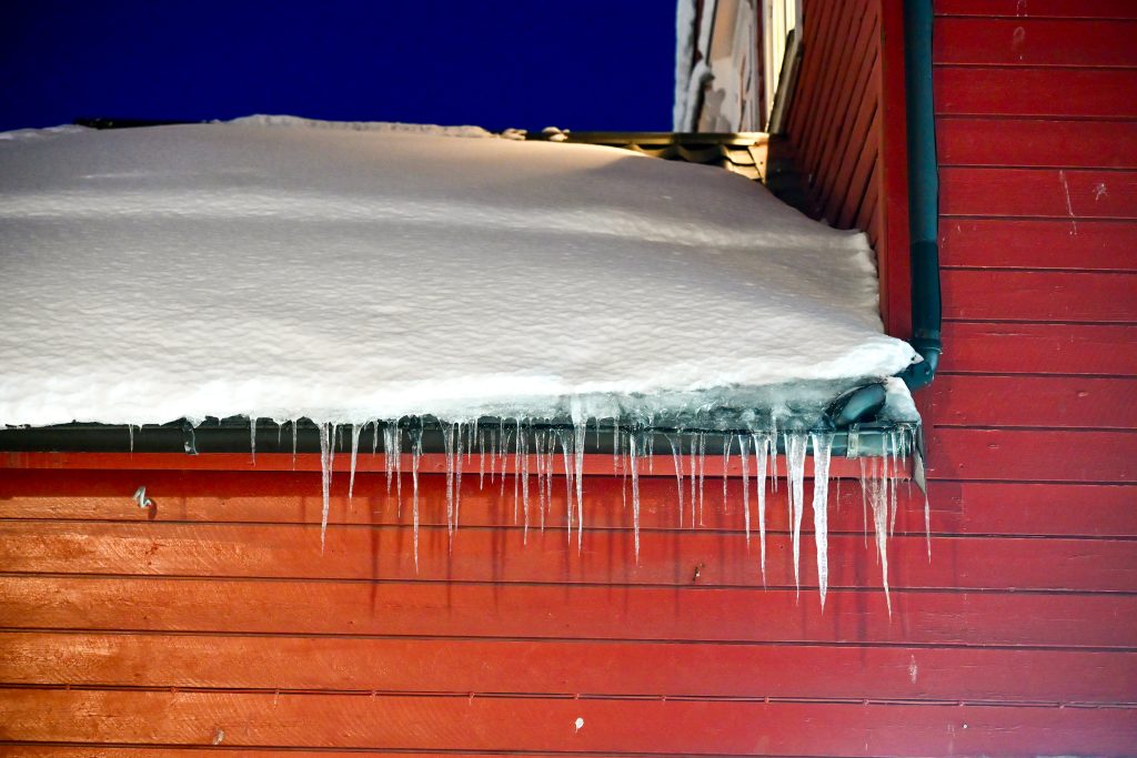 A row of icicles hangs from the gutter of a red wooden house with snow on the roof
