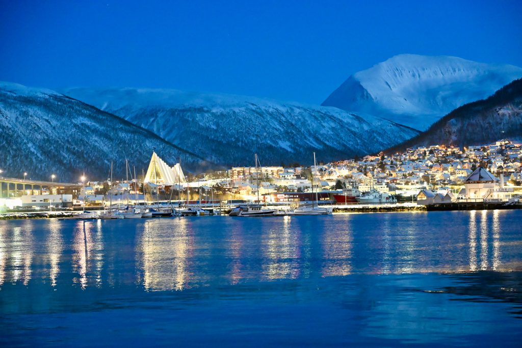 View at Twilight across water to land on the other side. The buildings on the shore are illuminated. A large church stands to the left and is reflected in the blue water. Mountains appear behind