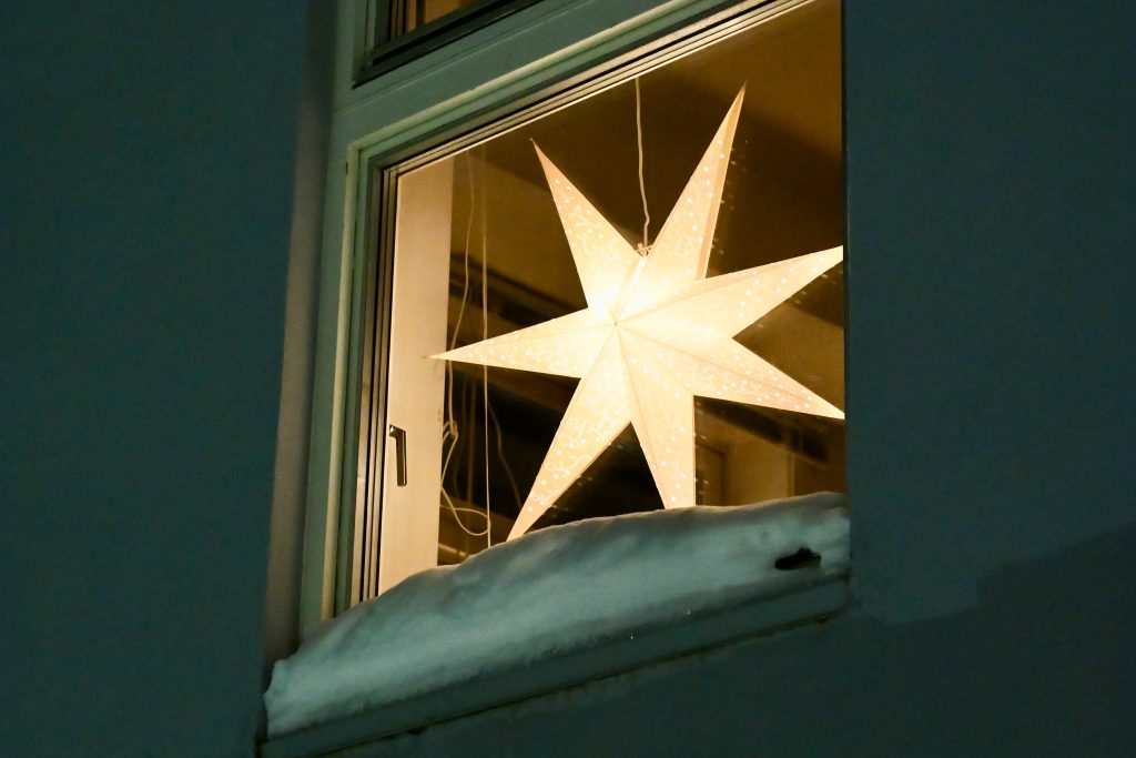 A window fills the frame with a single white paper star light in the middle. Dark green wooden walls surround