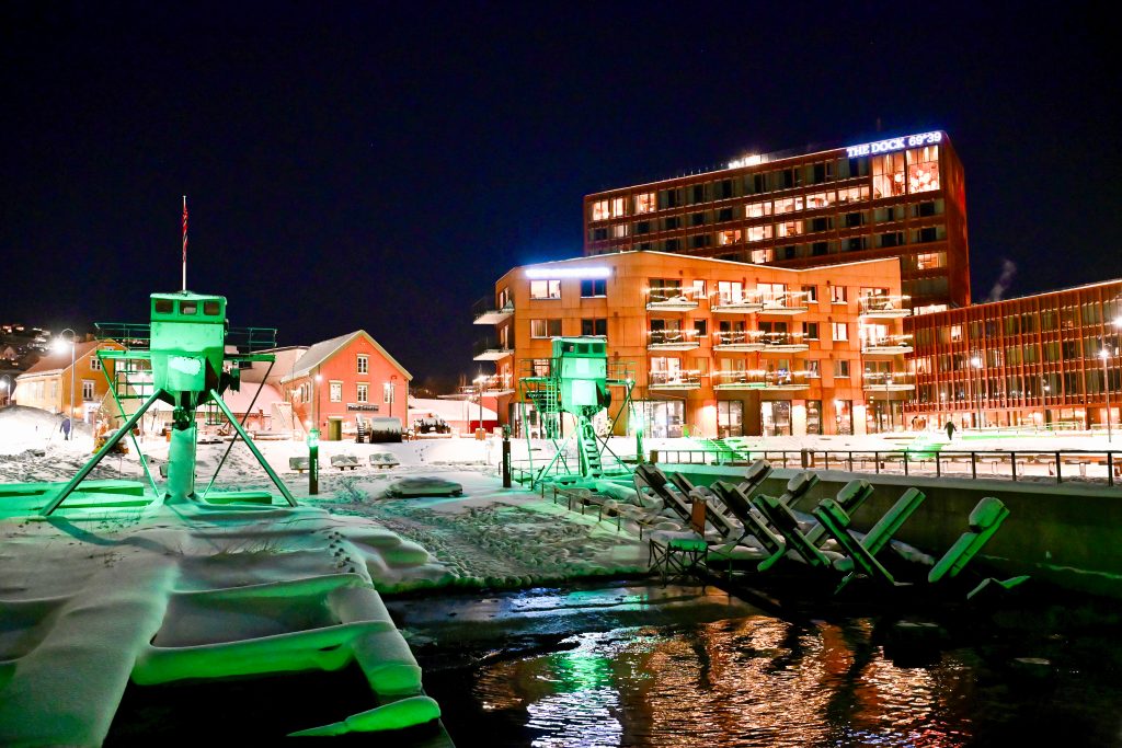 View across a small harbour at night towards some modern buildings behind, possibly hotels. There are green illuminated watch stations on the harbour