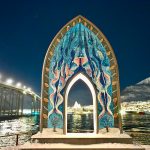 Dark night sky. A tall arched sculpture stands in the middle of the frame. It is covered in blue mosaic tiles. There is a whole in the middle through which you can see a white illuminated Church structure in the distance across a body of water. Lights of a bridge to the left