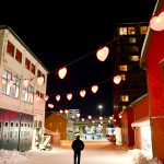 Dark sky above a snowy street. White wooden building to the left and red wooden building to the right. Strings of red heart lights criss cross the building. A solitary figure is walking away from the camera