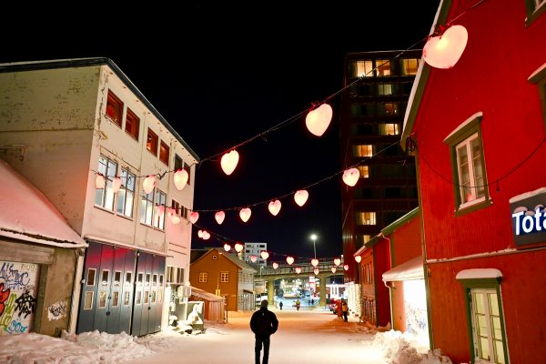 Dark sky above a snowy street. White wooden building to the left and red wooden building to the right. Strings of red heart lights criss cross the building. A solitary figure is walking away from the camera