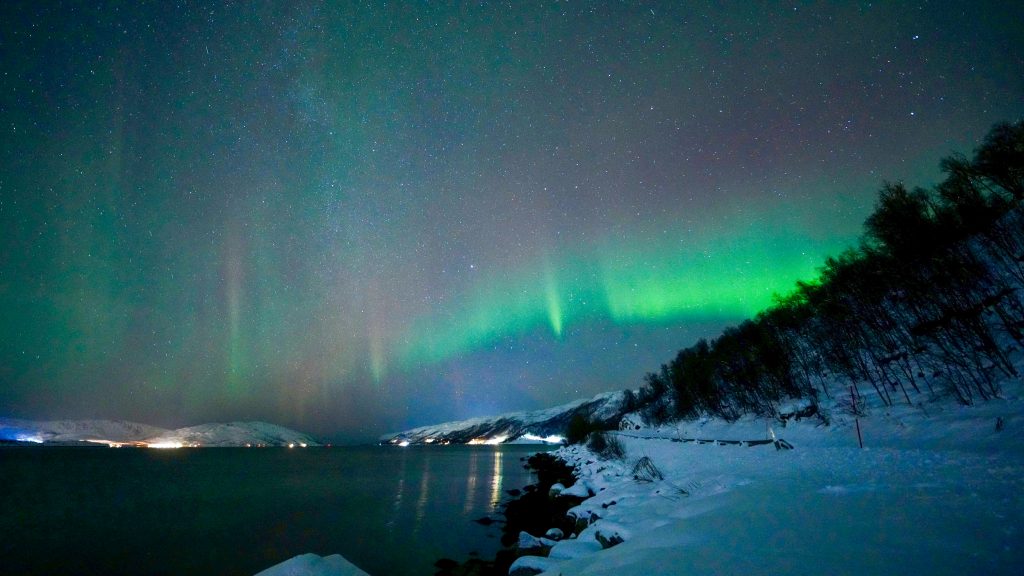 Two arcs of light run across the sky over a snowy landscape with a lake to the left.