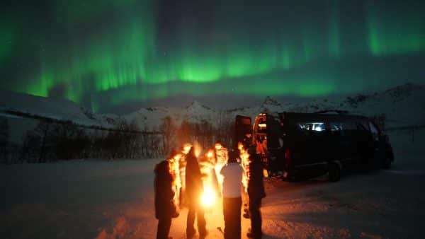 Night view of a group gathered around a campfire in the snow under green waves of Northern Lights