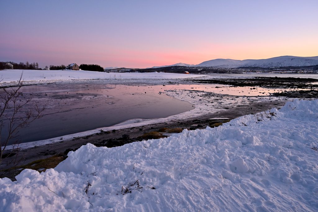 Gentle pink light fills the landscape sky and adds a pink glow to the still waters below. Snow in the foreground and on the horizon