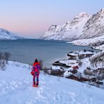 Beautiful snowy scene of a flat grey tear shaped lake or fjord surrounded by snowy mountains. Soft pink light on the horizon. Snow in front. A lady in a striking ski suit of bright pink stands in the centre looking at the water.