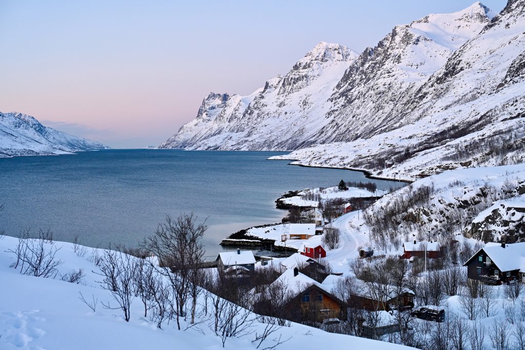 Landscape view from raised point in foreground looking across a body of water surrounded in a teardrop shape with snow covered mountains. Pinky glow in the sky above the water. Small number of village buildings in the foreground to the right