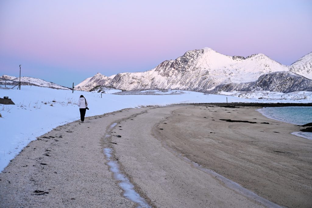Lanscape view. A lady beach curves around to the right from the bottom of the frame It is lined with snow, and there is a ridge of snow capped mountains on the horizon with pastel pink light above. A solitary figure walks away from the camera on the beach in white jacket and black trousers