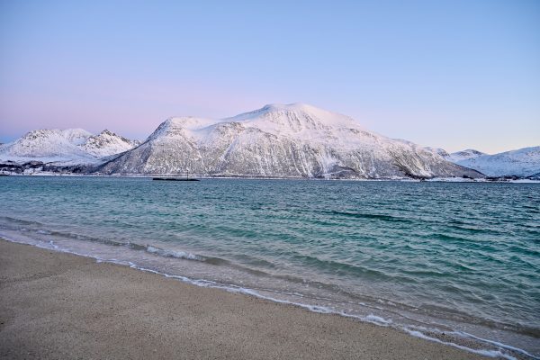View of a sandy beach diagonally against bottom left of frame, then tripe of turquoise water backed by snowy mountains and pale blue sky with a pink glow above