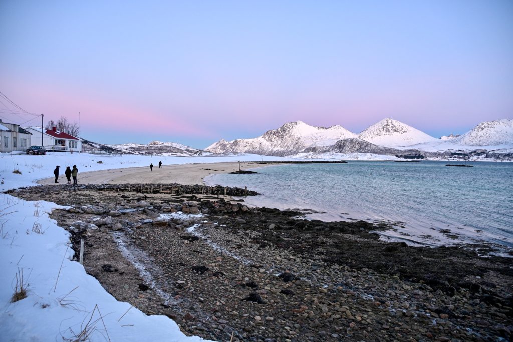 View across rocks to a sandy beach surrounded by snowy ground. Blue water to the right. White snow covered mountains in the background. Pink and blue light above the mountains