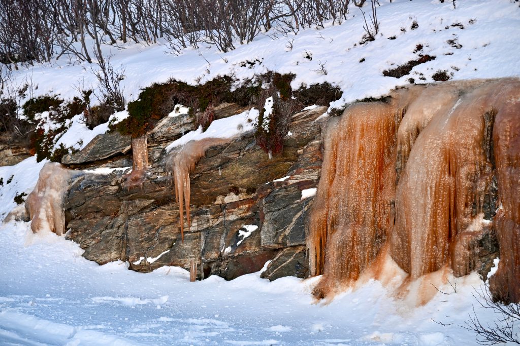 A stony wall fills the centre of the frame with frozen water falls across its length. Snow on the ground and on top of the wall