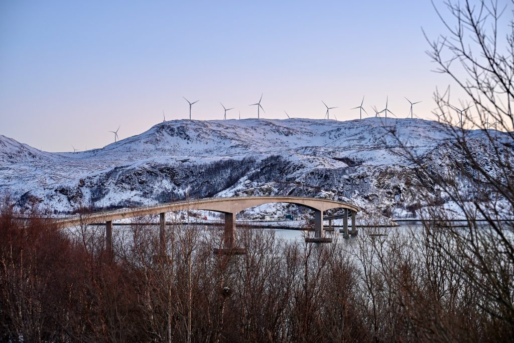 View over leafless shrubs along the bottom of the frame to body of water. A road bridge crosses the water. Snowy mountain at other side of the bridge. Row of wind turbines on the mountain