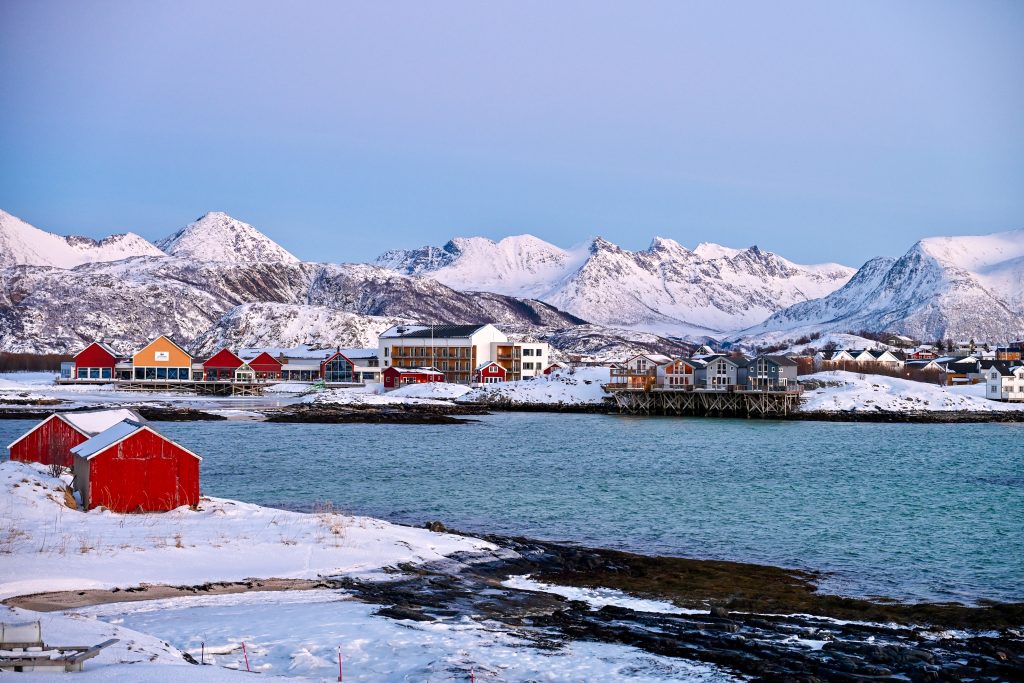 View across water to snowy mountains beyond. A number of wooden huts on stilts line the waterline and stand out agains the snow