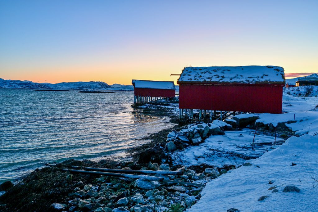 View from the ground in bottom right over water. Two red wooden cabins on stilts stand to the top right of the frame. Sunset to the left hand side horizon