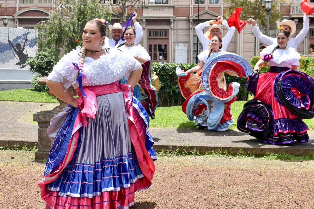 Lady stands in left of frame in colourful full skirt and white shirt, row of dancers behind. look to be wearing traditional outfits