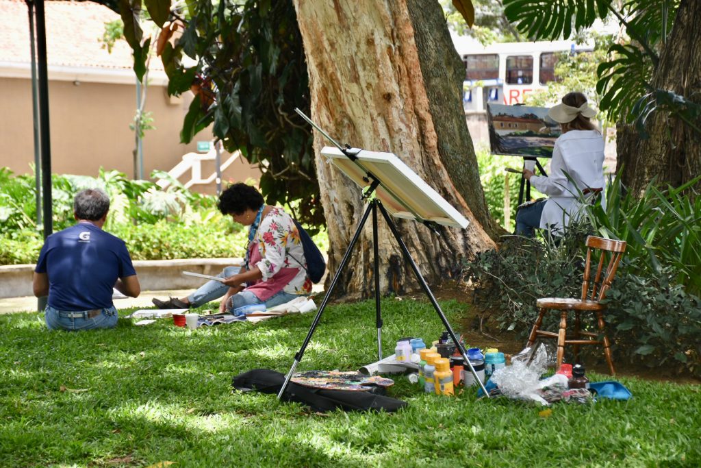 Park scene of grass in the front and a tree behind. Lady is painting at an easel. Lady is reading under a tree
