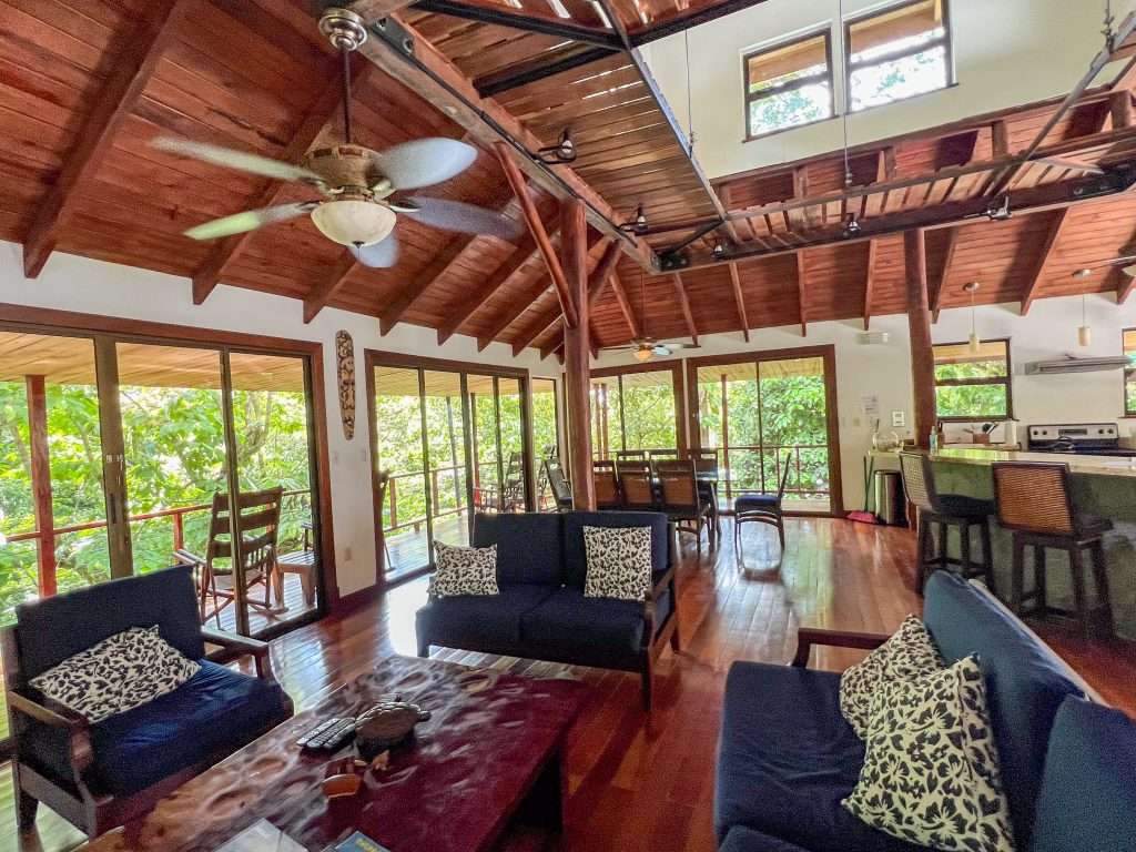 View of the inside of a lounge I a wooden. building with large windows to the left and far side. A blue sofa sits in the middle of the room and chairs around the outside