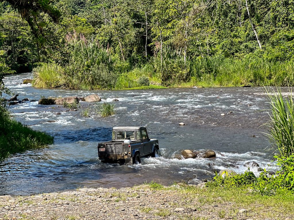 Grey Land Rover crossing to the far side of a river with trees on other side