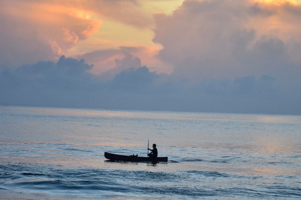 A boat in silhouette on a still sea with peachy sunset clouds behind