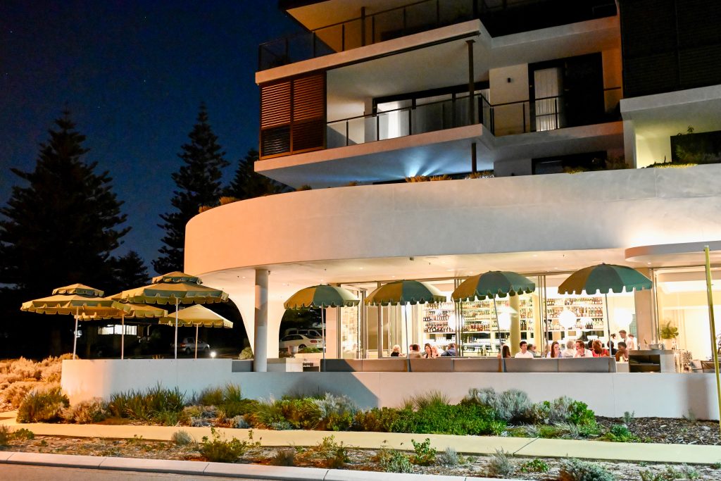 View of an Art Deco style restaurant at night. It has a curved white roof on the first floor with more storey above. Road and grass in front. Lights are on, people inside the restaurant