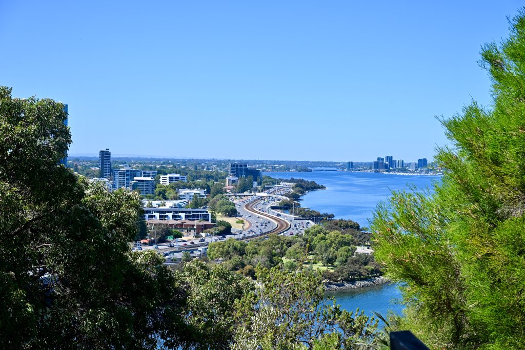 Looking down on a river winding in front of the frame and to the right hand side. A windy road, buildings and trees to the left. Pale sky above