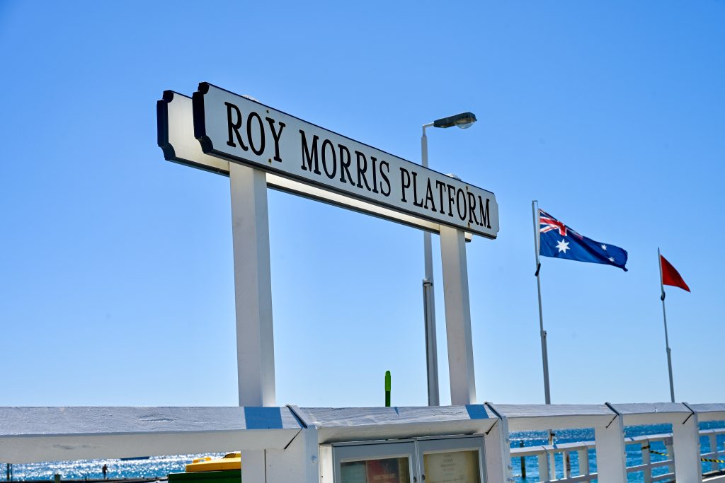White wooden sign saying 'Road Morris Platform' with the sea and Australian flag in the background. Blue sky