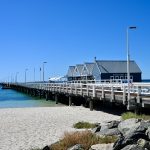 View across golden sand to a white wooden pier running from the right of the photo across to the horizon on the left. A row of wooden huts sits on the pier. Blue sky above. Turquoise sea visible to the left