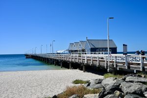 View across golden sand to a white wooden pier running from the right of the photo across to the horizon on the left. A row of wooden huts sits on the pier. Blue sky above. Turquoise sea visible to the left
